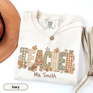 May include: An ivory t-shirt featuring the word TEACHER in large, patterned block letters with autumn-themed designs including plaid, floral, and geometric prints. Below the text, the name Ms. Smith is printed in brown, alongside small illustrations of books, an apple, and a pencil.