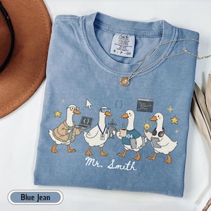May include: A blue jean colored t-shirt featuring a graphic of four white ducks dressed as tech workers. The ducks are depicted with a laptop, a robot, a coffee mug, and a lightbulb. The text "Mr. Smith" is printed below the ducks in white script.