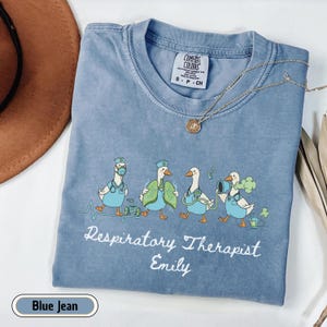 May include: Blue jean colored t-shirt with a graphic of cartoon geese dressed as respiratory therapists. The text "Respiratory Therapist Emily" is printed below the geese. A silver necklace with a pendant is on the shirt.