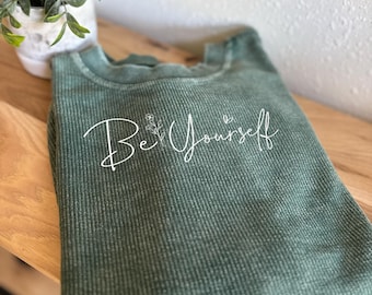 May include: A green corded sweatshirt with the words "Be Yourself" in white script. The text is accompanied by a small floral design. The sweatshirt is folded on a wooden surface, with a small potted plant in the background.