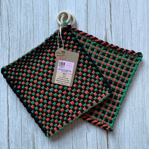 May include: Two black, green, and orange woven pot holders with a wooden ring handle. The pot holders are heat resistant and washable. The tag reads "Hand made" and "Our handmade pot holders are heat resistant and washable, but hang to dry."