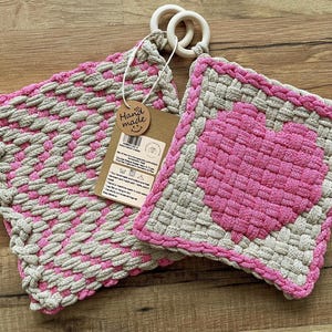 May include: Two handmade crocheted pot holders, one with a pink heart design and the other with a pink and beige checkered pattern. The pot holders are connected by a wooden ring. The pot holders have a "Hand Made" tag attached.