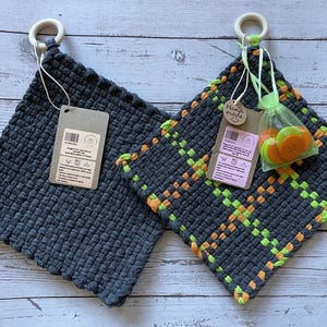 May include: Two handmade woven pot holders with a gray background and colorful patterns. One pot holder has a green and orange pattern, the other has a solid gray color. Both pot holders have a white ring attached to the top.