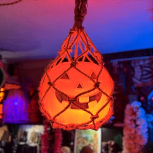 May include: A glowing orange pumpkin-shaped Halloween decoration with a carved face, suspended from the ceiling in a macrame net. The pumpkin is lit from within, creating a warm, inviting glow. The rope is a natural brown color.