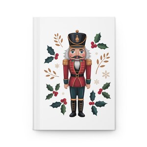 Puede incluir: Un diario blanco con una ilustración colorida de un soldado cascanueces. El cascanueces viste un abrigo rojo, botas negras y un sombrero negro. Acebo, bayas, copos de nieve y ramitas adornan el diseño.