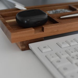 May include: A wooden desk organizer with a black wireless earbud case, a silver pen, and a white keyboard. The organizer has compartments for small items and is made of a warm-toned wood.