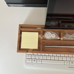 May include: A wooden desk organizer with compartments for office supplies. It holds a yellow sticky note, a white correction tape dispenser, paper clips, and a pen. A white keyboard is below the organizer. The monitor above has "HP Eye Ease" printed on it.