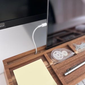 May include: A wooden desk organizer with compartments for office supplies. Includes a yellow sticky note pad, a white correction tape dispenser, a silver pen, and a white keyboard. A computer monitor is in the background.