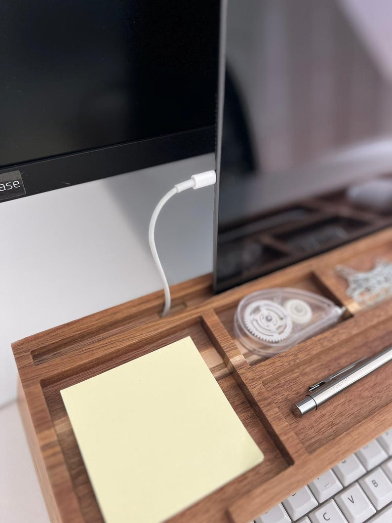 May include: A wooden desk organizer with compartments for office supplies. Includes a yellow sticky note pad, a white correction tape dispenser, a silver pen, and a white keyboard. A computer monitor is in the background.