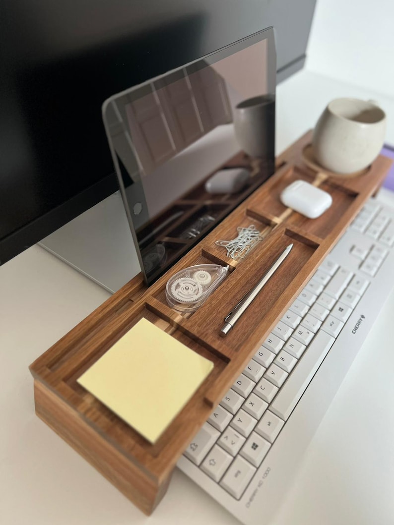 May include: A wooden desk organizer with compartments for a tablet, a mug, a pen, and other office supplies. A white keyboard is positioned below the organizer. A yellow sticky note pad is visible. The organizer is made of dark wood.