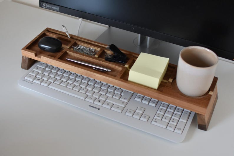 May include: Wooden desk organizer with compartments for office supplies, including a pen, paper clips, and a stapler. A black computer mouse, a notepad, and a ceramic mug are also visible. The organizer sits above a keyboard.