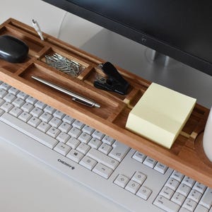 May include: A wooden desk organizer with compartments for office supplies, including a pen, paperclips, and a stapler. A black mouse, sticky notes, and a ceramic mug are also visible. The organizer sits above a white keyboard.