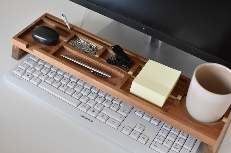 May include: A wooden desk organizer with compartments for office supplies, including a pen, paperclips, and a stapler. A black mouse, sticky notes, and a ceramic mug are also visible. The organizer sits above a white keyboard.
