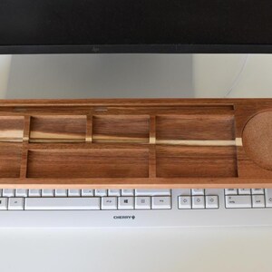 May include: A wooden keyboard tray with multiple compartments and a built-in coaster. The tray is made of dark brown wood with lighter wood accents and sits above a white keyboard. The tray is designed to hold office supplies.