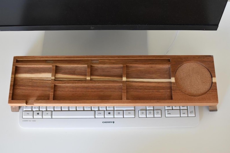 May include: A wooden keyboard tray with multiple compartments and a built-in coaster. The tray is made of dark brown wood with lighter wood accents and sits above a white keyboard. The tray is designed to hold office supplies.
