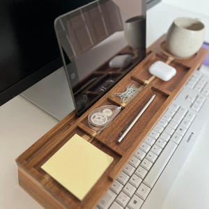 May include: A wooden desk organizer with compartments for a tablet, a mug, a pen, and other office supplies. A white keyboard is positioned below the organizer. A yellow sticky note pad is visible. The organizer is made of dark wood.
