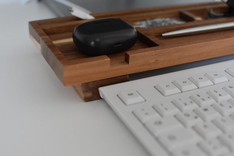 May include: A wooden desk organizer with a black wireless earbud case, a silver pen, and a white keyboard. The organizer has compartments for small items and is made of a warm-toned wood.