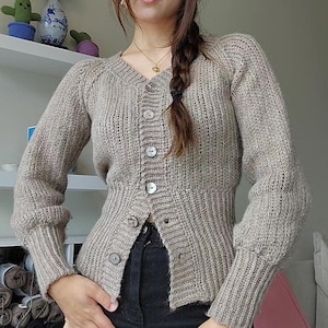 Op de afbeelding: Beige vest met knopen, een V-hals en geribbelde mouwen. De gebreide trui heeft een getailleerde taille en wordt gedragen met een zwarte broek. Het vest heeft grote, ronde knopen.