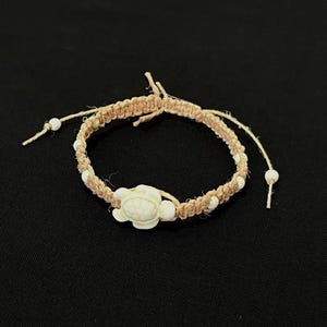 May include: A tan woven cord bracelet featuring a central white turtle charm and small white spherical beads integrated into the band. The bracelet has adjustable knotted ends with white beads at the tips, creating a flexible and secure fit.