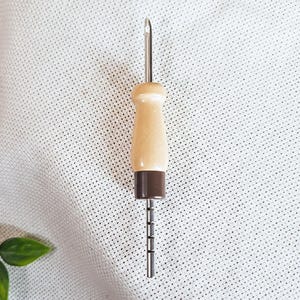 Può includere: Uno strumento per punch needle con un manico in legno marrone chiaro e una base marrone scuro. L'ago in metallo argentato ha un piccolo occhiello sulla punta. Questo strumento è progettato per il rug hooking e il ricamo.