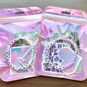 May include: Two clear plastic bags with zippers, each containing a white beaded bracelet with black letter beads spelling out song titles. The left bag has a bracelet that says "THE MANUSCRIPT" and the right bag has a bracelet that says "LAVENDER HAZE".