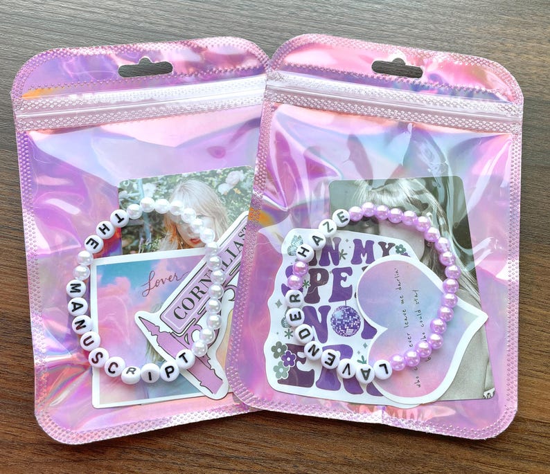 May include: Two clear plastic bags with zippers, each containing a white beaded bracelet with black letter beads spelling out song titles. The left bag has a bracelet that says "THE MANUSCRIPT" and the right bag has a bracelet that says "LAVENDER HAZE".