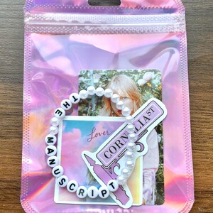 May include: A white pearl bracelet with black letter beads spelling "The Manuscript" and a sticker with the text "Cornelia St" and "Lover" on a pink background.
