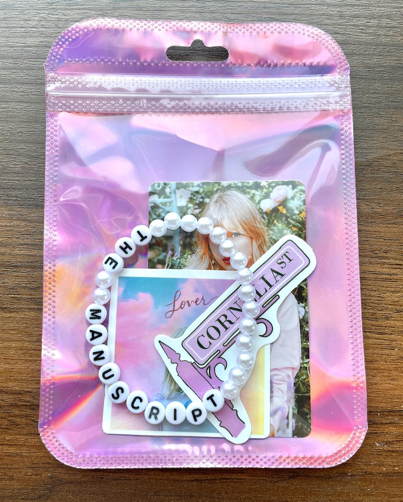 May include: A white pearl bracelet with black letter beads spelling "The Manuscript" and a sticker with the text "Cornelia St" and "Lover" on a pink background.