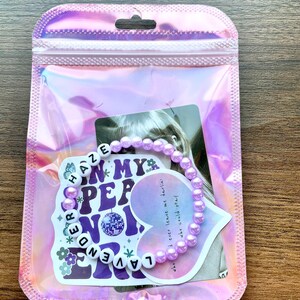 May include: A purple beaded bracelet with the words "Lavender Haze" and "In My  Pep No" on a white heart-shaped sticker. The sticker also includes the lyrics "who I ever leave me darlin' who could stay".