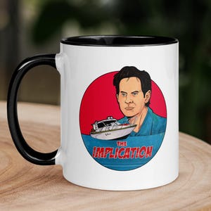 Op de afbeelding: Witte keramische mok met een zwarte handgreep en binnenkant. De mok heeft een cirkelvormig ontwerp met een man, een boot en de tekst "THE IMPLICATION" in blauw en rood. De mok staat op een houten oppervlak.