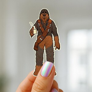 May include: A sticker featuring a cartoon illustration of Chewbacca holding a mug. The character is depicted in his signature brown fur with a bandolier. The sticker is held up by a hand with iridescent nail polish and gold rings.