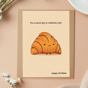Peut inclure: Carte de v&oelig;ux d'anniversaire avec une illustration de croissant souriant. Le texte "It's a sweet day to celebrate you!" est en haut et "happy birthday" en bas. Le fond est neutre, avec un cadre marron.