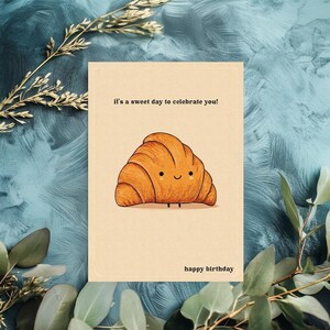 Peut inclure: Une carte d'anniversaire beige avec une illustration de croissant de dessin anim&eacute;. Le croissant orange a un visage souriant et de petites jambes. Le texte dit "it's a sweet day to celebrate you!" et "happy birthday".