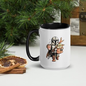 Op de afbeelding: Witte keramische mok met een zwarte binnenkant en handvat. De mok heeft een cartoon illustratie van een personage met een helm die een kopje vasthoudt, met een klein groen wezen met een jasje. De mok heeft een inhoud van ongeveer 325 ml.