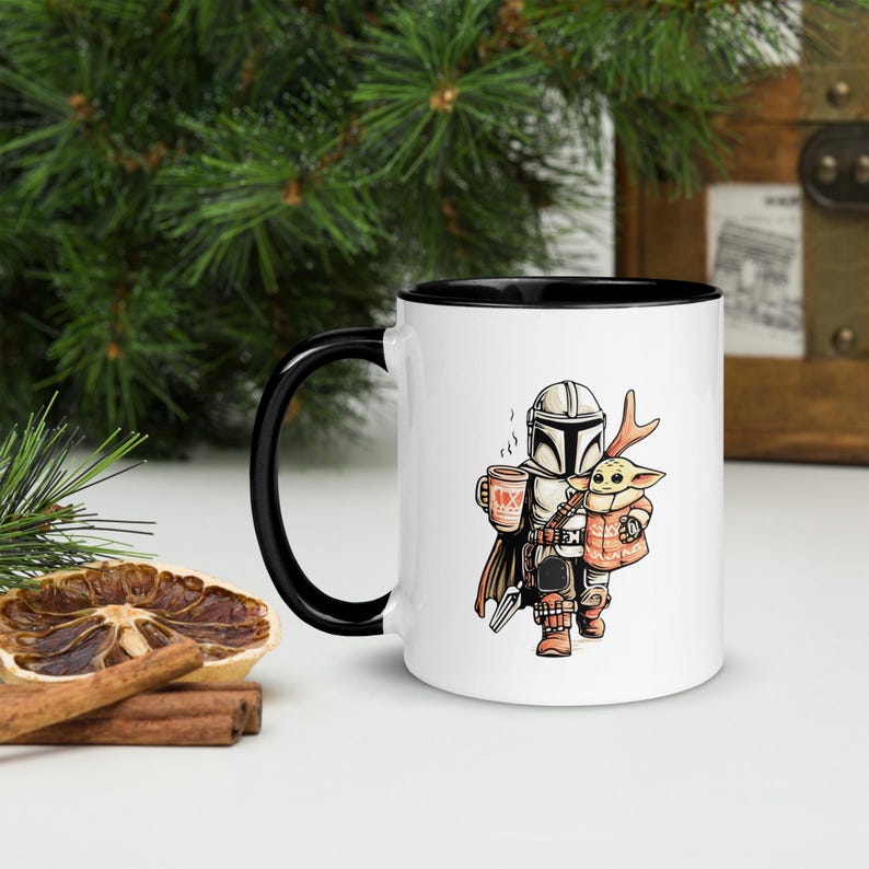 Op de afbeelding: Witte keramische mok met een zwarte binnenkant en handvat. De mok heeft een cartoonillustratie van een personage met een helm die een beker met een hart erop en een klein groen wezen vasthoudt. De mok staat op een wit oppervlak.