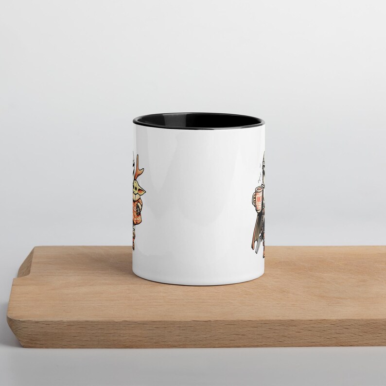 Op de afbeelding: Witte keramische mok met een zwarte binnenkant en twee cartoon illustraties. E&eacute;n kant toont een personage met grote oren en een bruin outfit, de andere kant toont een personage in een zwarte cape met een mok. De mok staat op een houten oppervlak.