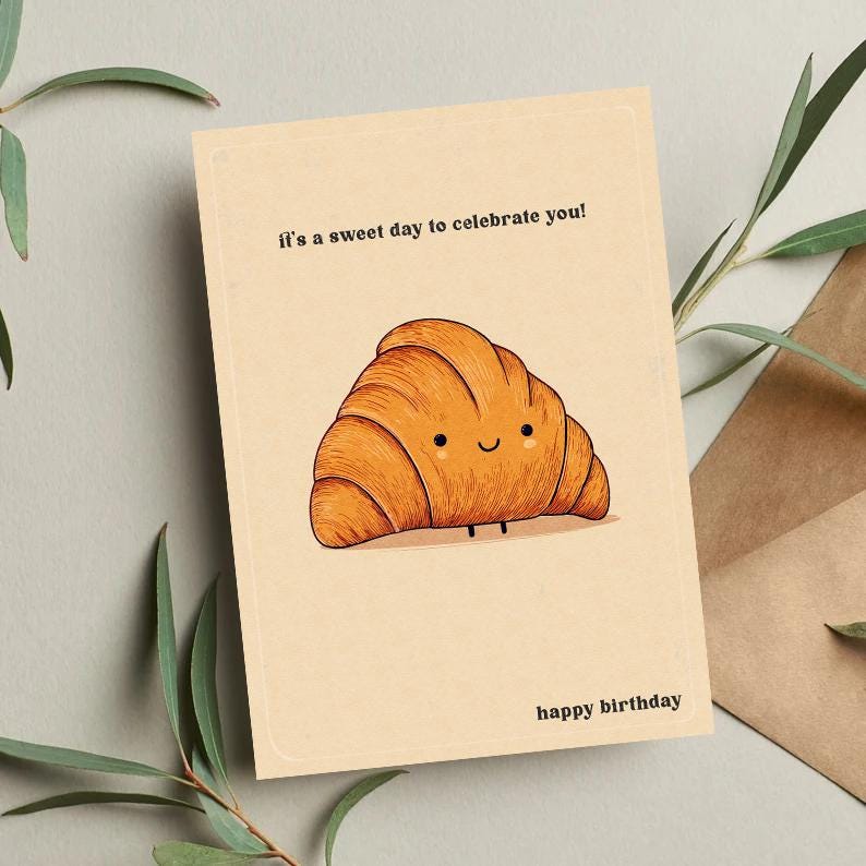Peut inclure: Carte de v&oelig;ux beige avec une illustration de croissant de dessin anim&eacute;. Le texte "It's a sweet day to celebrate you!" est en haut et "happy birthday" en bas. Le croissant a un visage souriant.