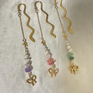 May include: Three gold-coloured metal bookmarks with a wavy design. Each bookmark has a chain with beads and a gold bow charm. The beads are various colours, including purple, pink, and green. These are ideal for book lovers.