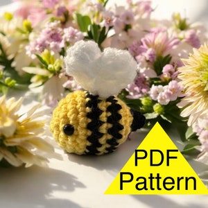May include: A crocheted bee with yellow and black stripes, a white heart-shaped cloud, and black eyes. The bee is set against a backdrop of flowers. A yellow triangle with the text "PDF Pattern" is in the lower right corner.