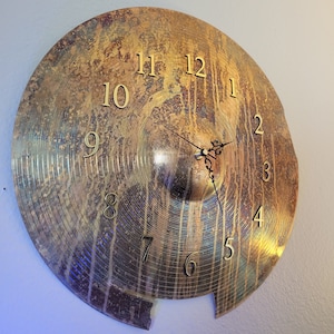 Op de afbeelding: Een gouden cymbal-klok met Romeinse cijfers. De cymbal heeft een verweerde, oude uitstraling met een gebroken rand.