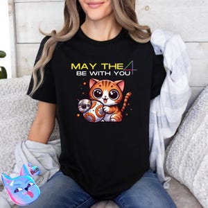 以下が含まれることがあります： BB-8 ドロイドを持った猫の白と黄色のグラフィックが描かれた黒いTシャツ。Tシャツには「May the 4 be with you」という文字が印刷されています。