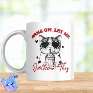 Puede incluir: Taza de cerámica blanca con una ilustración en blanco y negro de un gato con gafas de sol en forma de corazón y bebiendo un batido. El texto "Hang on, let me overthink this" está escrito en rojo en la taza.