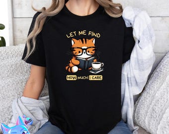 May include: Black t-shirt featuring a cartoon tiger wearing glasses and reading a book, with a coffee cup and the text "LET ME FIND HOW MUCH I CARE". The tiger is orange and brown. The shirt is a casual wear item.