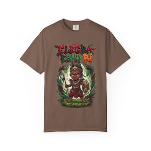 May include: Brown t-shirt featuring a colorful graphic of a warrior with a spear and shield, surrounded by foliage. The text "Elegba Eshu Bi" is displayed above the warrior in a graffiti-style font. The shirt is made of a soft material.