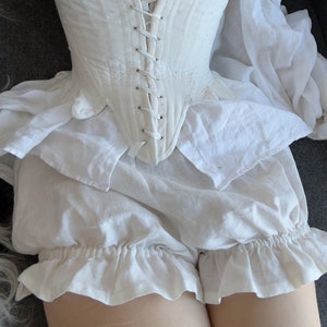 May include: A white cotton corset with a lace-up front and a ruffled white skirt. The corset has a fitted bodice and a boned structure.