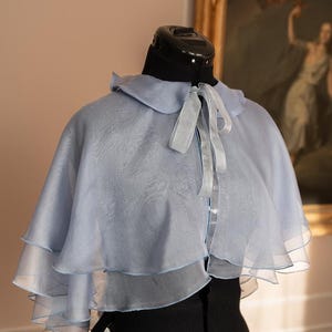 Puede incluir: Una capa azul claro de varias capas con cuello redondo y lazo de cinta de raso. La capa se exhibe en un maniquí negro. Un cuadro en un marco dorado se encuentra en el fondo.