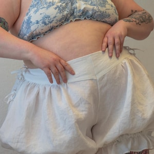 May include: White cotton bloomers with a ruffled trim. The bloomers have a wide waistband and are gathered at the legs.