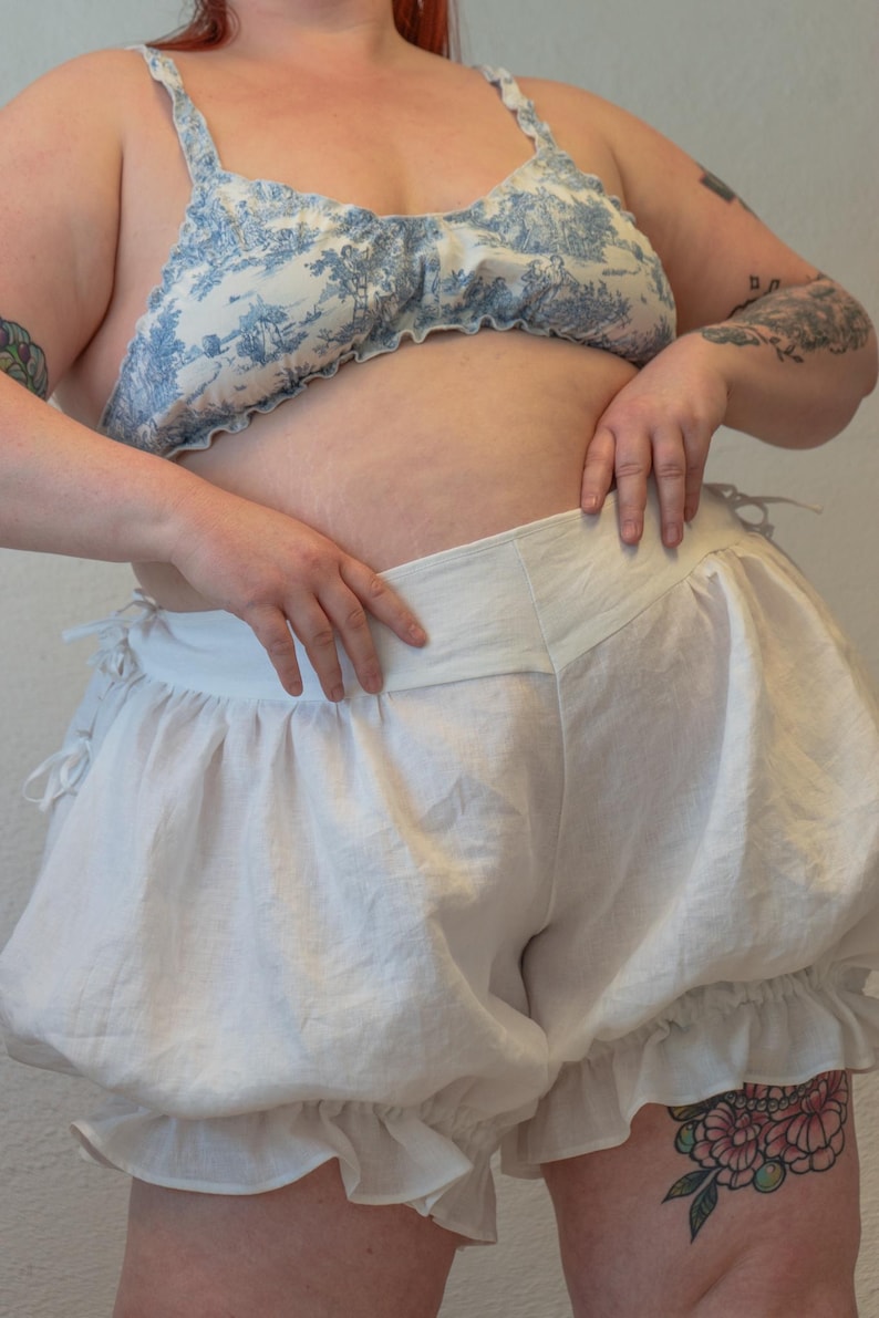 May include: White cotton bloomers with a ruffled trim. The bloomers have a wide waistband and are gathered at the legs.