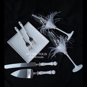 May include: White wedding cake knife and server with white handles and a silver blade. Two white forks are on a white square plate. Two champagne flutes with white feathers and rhinestones are on a black background. The text "STUDIO FOLIS handmade" is on the plate.