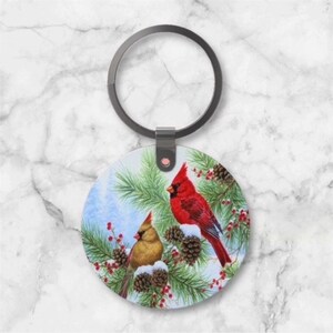 May include: A round keychain featuring a winter scene with two cardinals perched on snow-covered pine branches. The keychain has a silver-colored ring and a clasp.